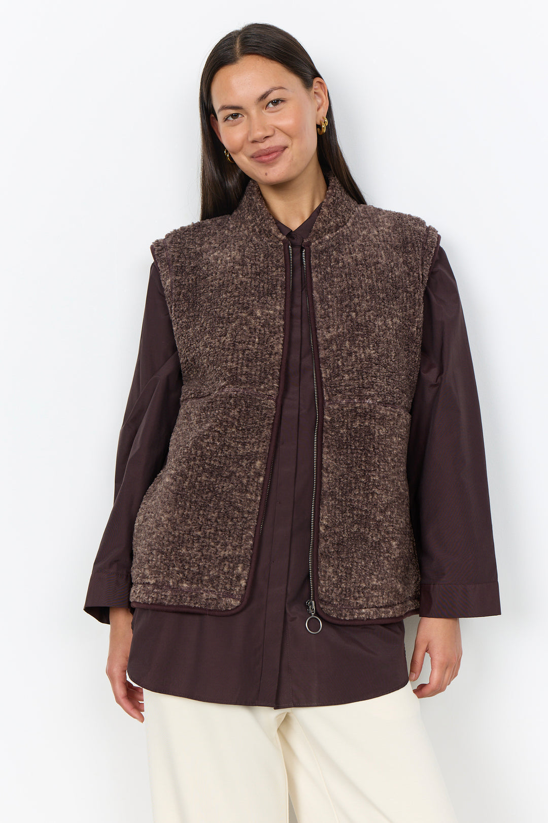 A woman modelling a brown boucle vest and cream pants in front of a white background. 