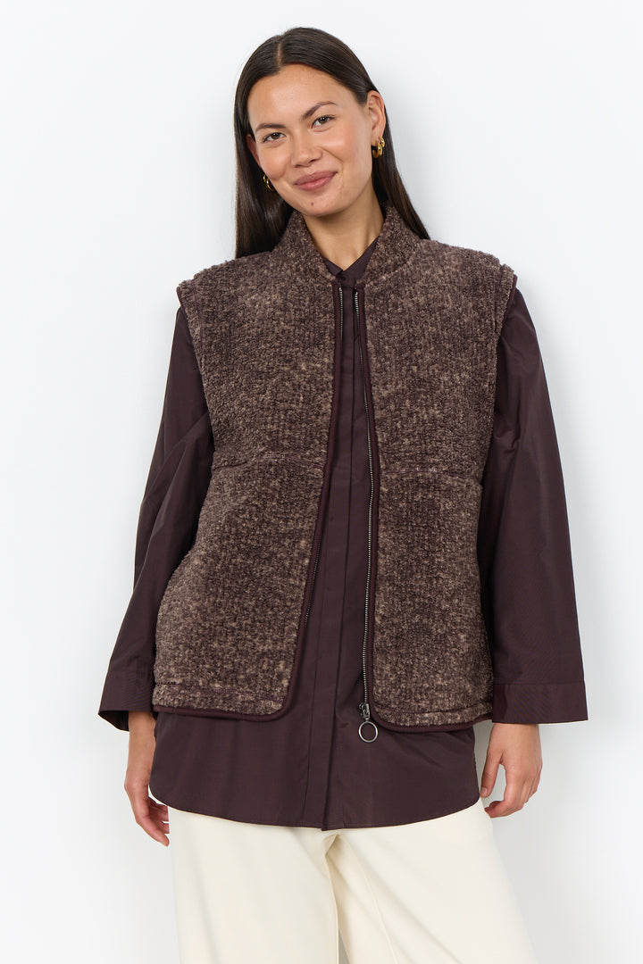A woman modelling a brown boucle vest and cream pants in front of a white background. 
