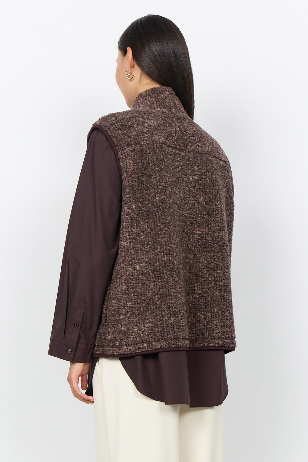 A woman modelling a brown boucle vest and cream pants in front of a white background. 