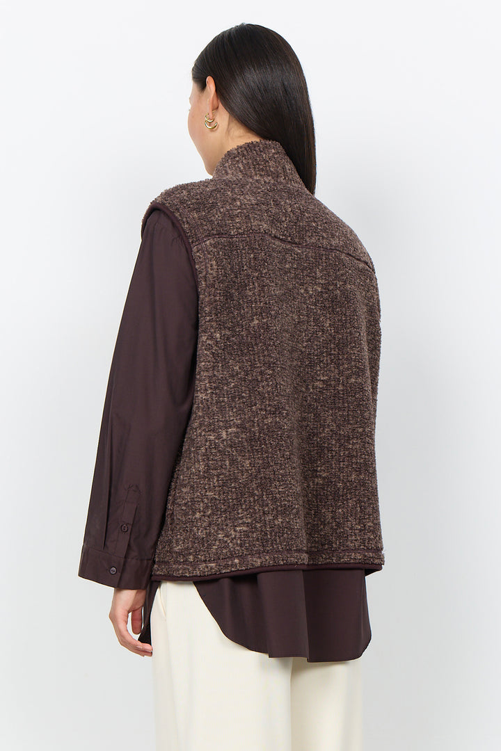 A woman modelling a brown boucle vest and cream pants in front of a white background. 