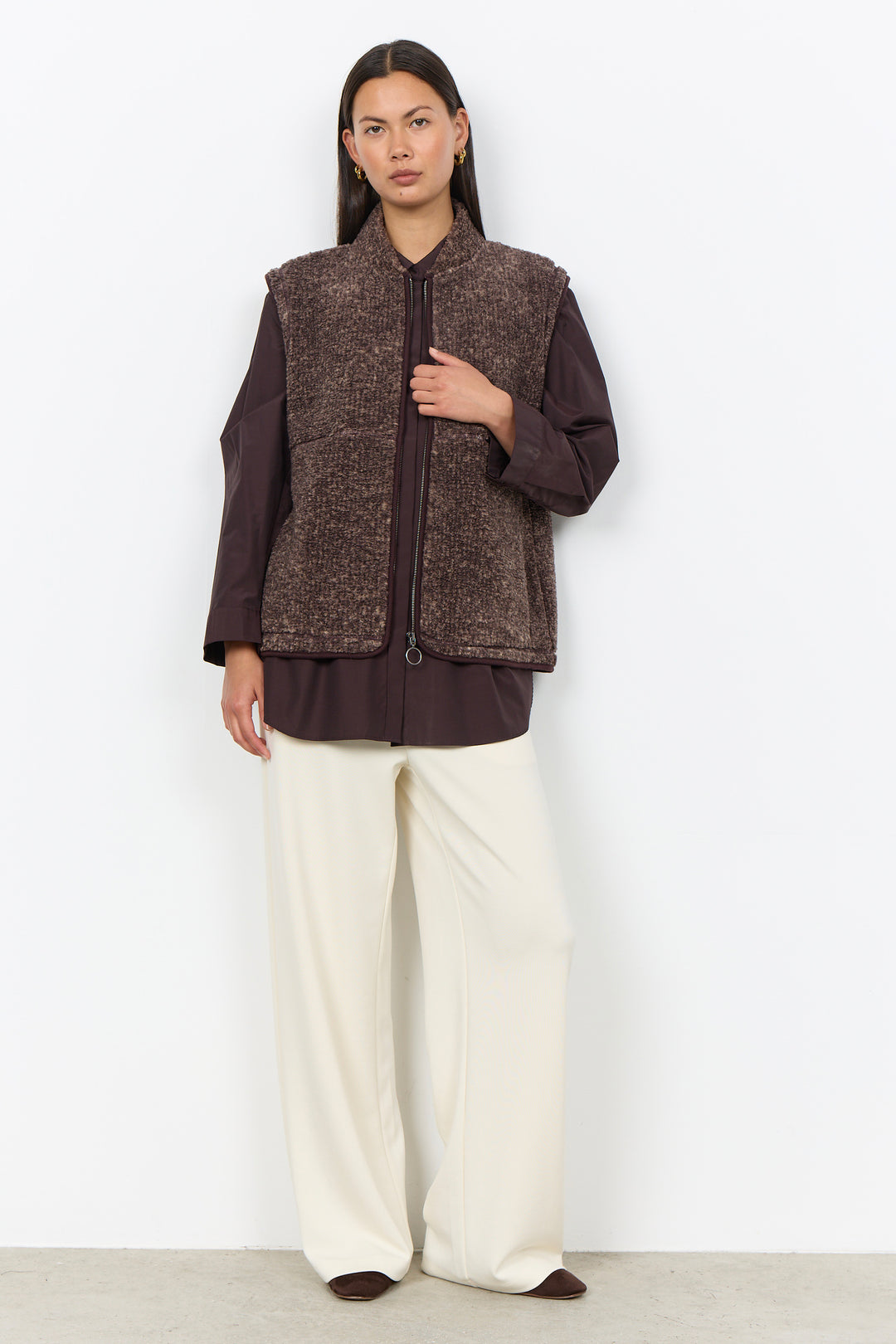 A woman modelling a brown boucle vest and cream pants in front of a white background. 