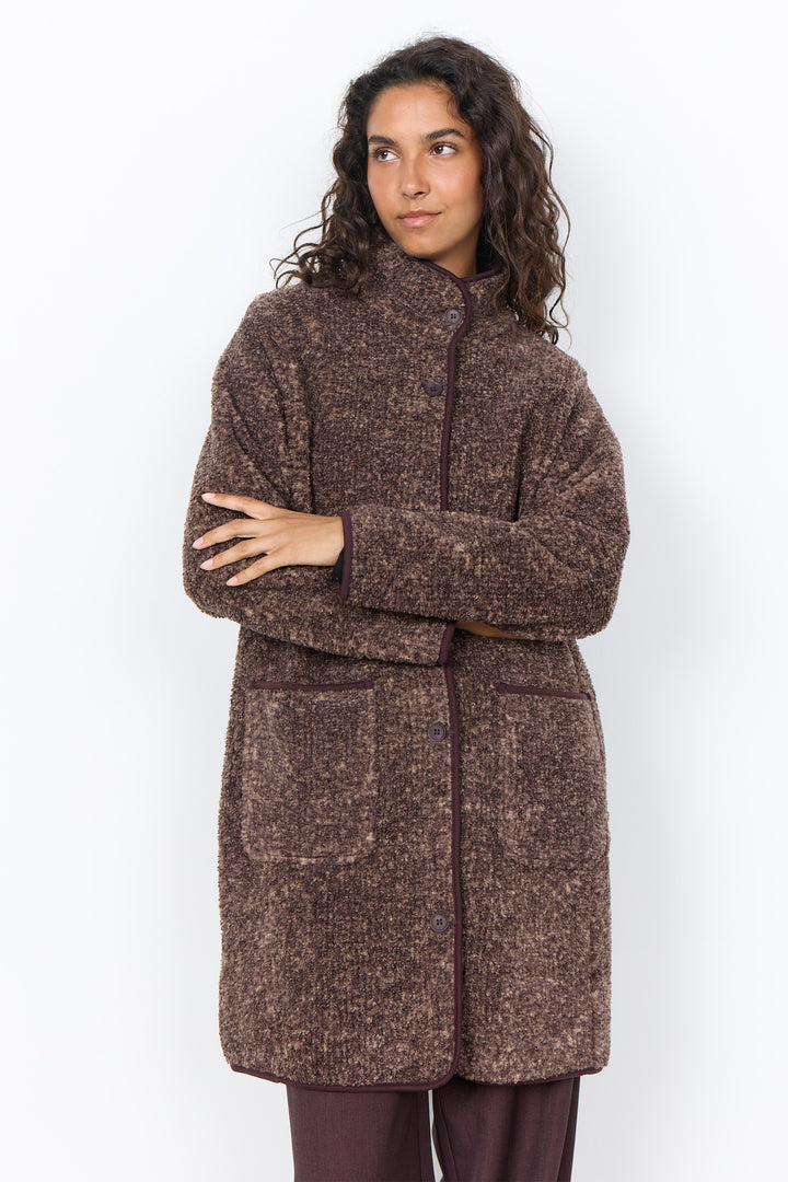 A woman modelling a long brown boucle coat with brown pants in front of a white background. 