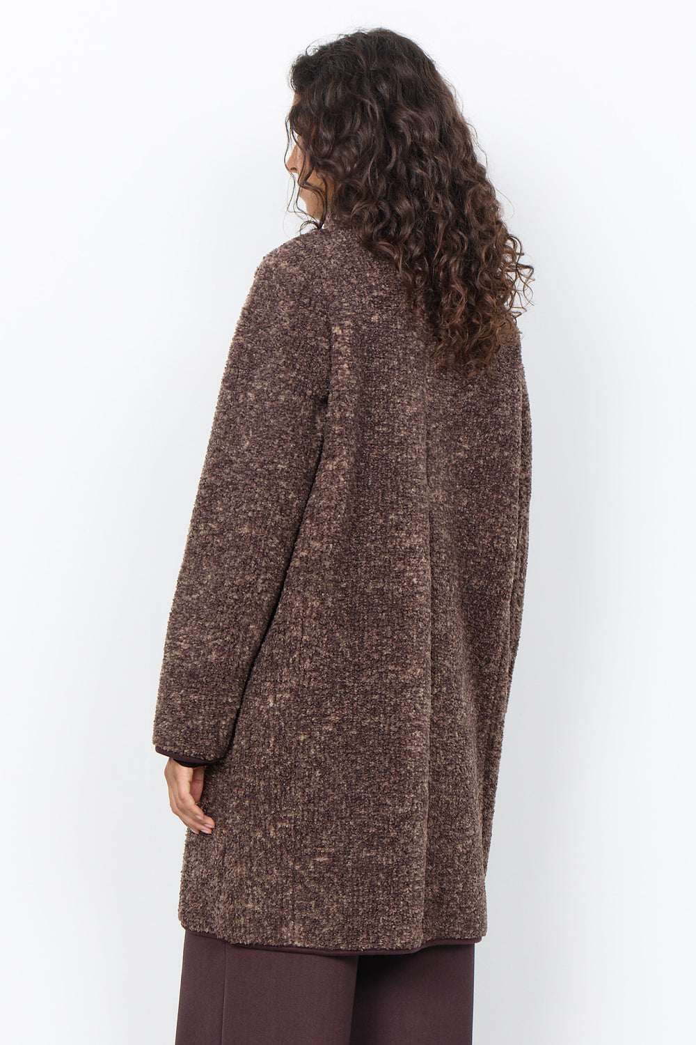 A woman modelling a long brown boucle coat with brown pants in front of a white background. 