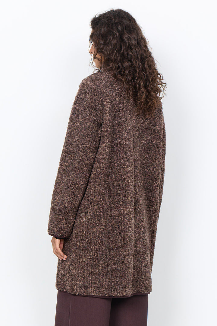A woman modelling a long brown boucle coat with brown pants in front of a white background. 