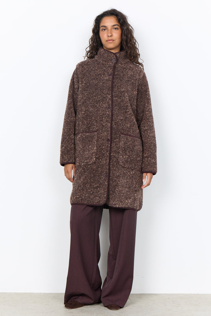 A woman modelling a long brown boucle coat with brown pants in front of a white background. 