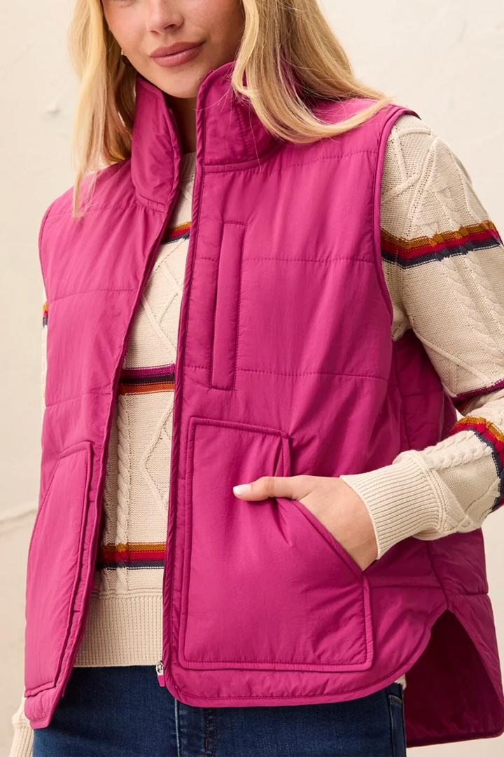 A woman modelling a pink puffer vest and sweater standing in front of a light beige background. 