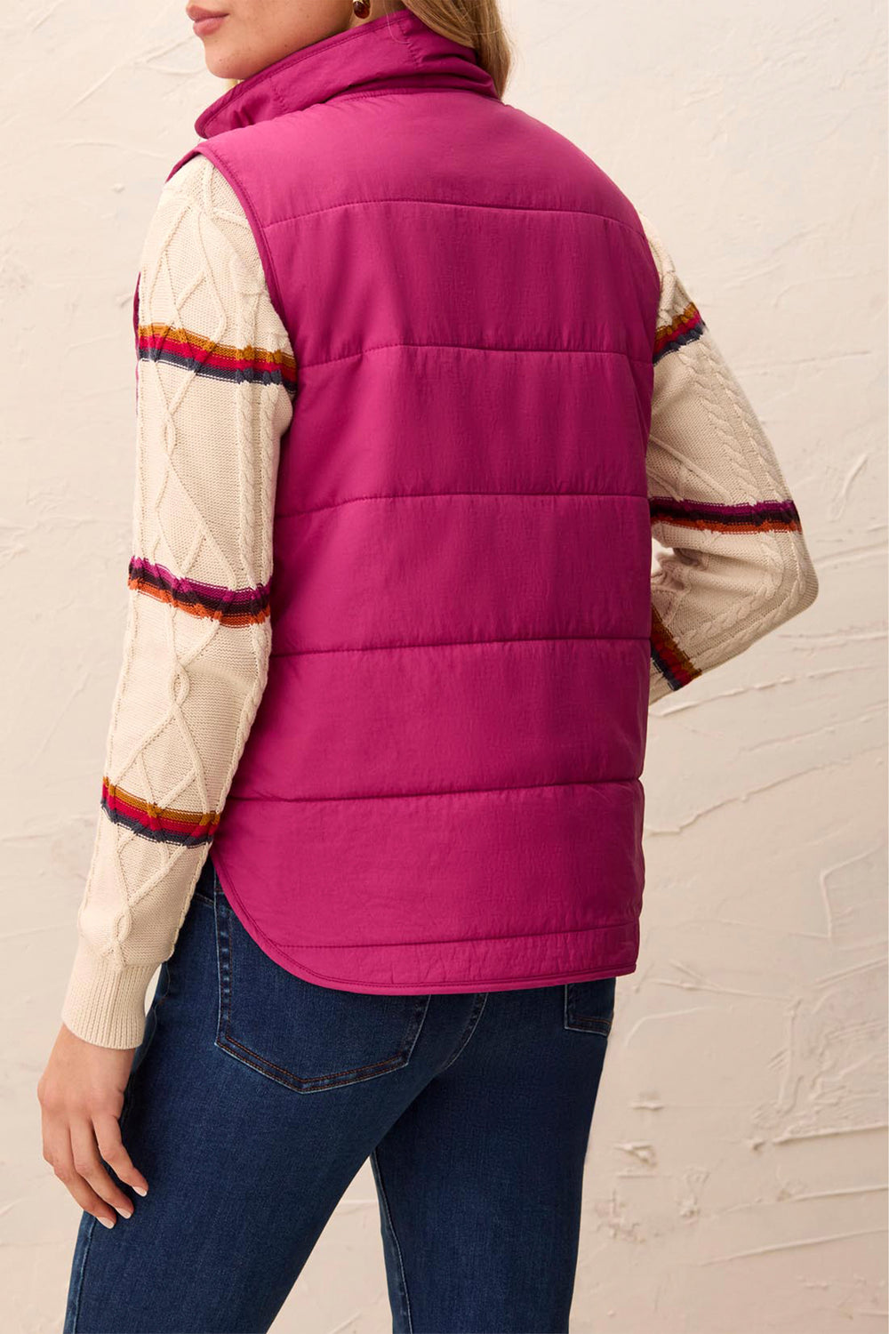 A woman shown from the back modelling a pink puffer vest and sweater standing in front of a light beige background. 