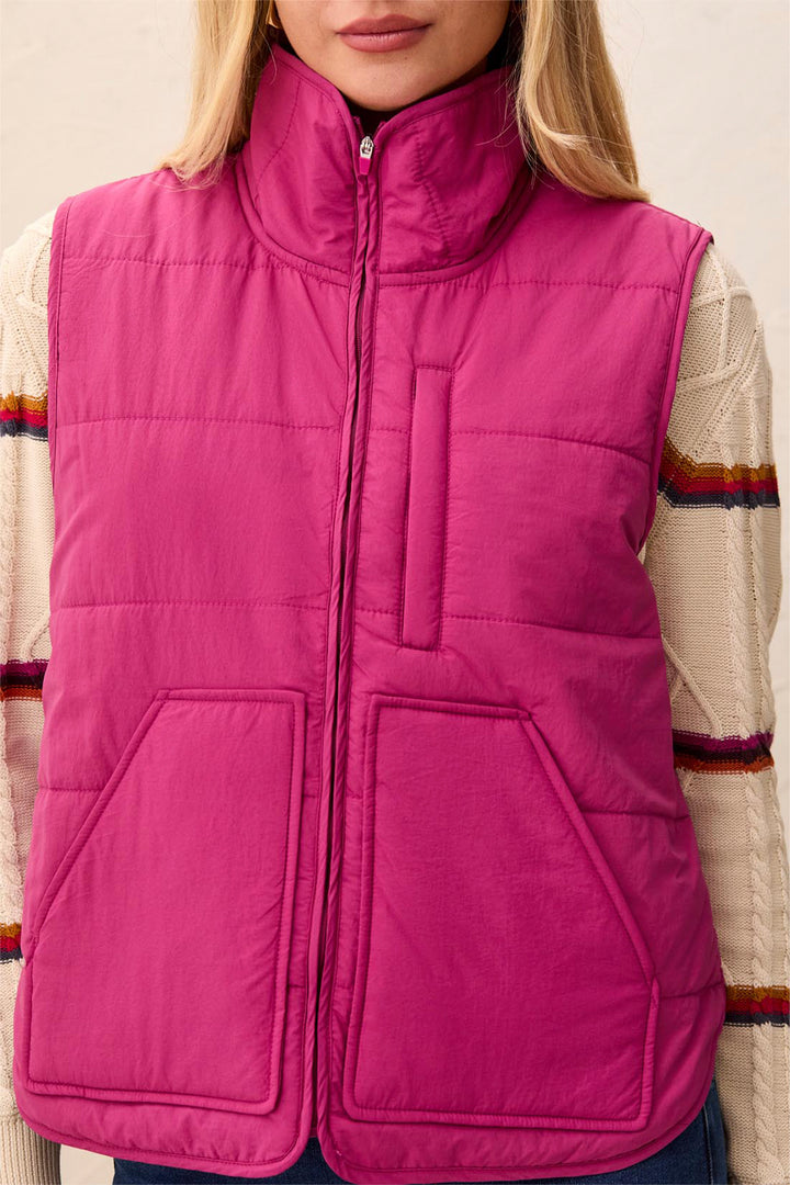 A woman modelling a pink puffer vest and sweater standing in front of a light beige background. 