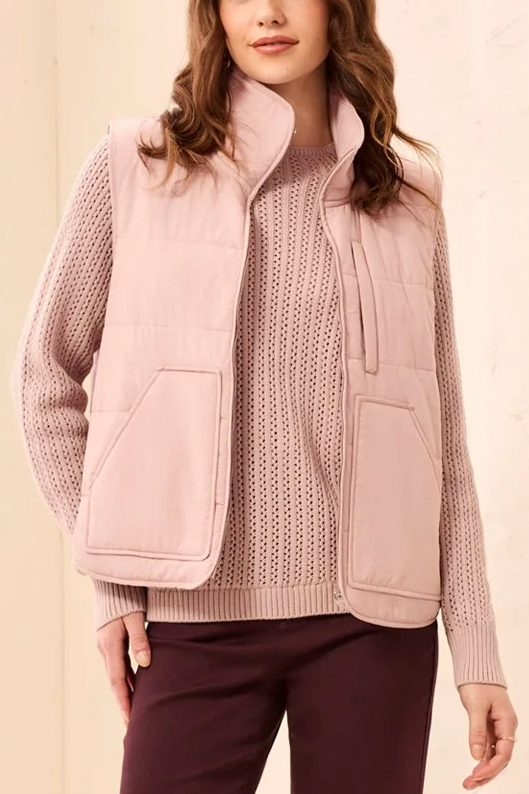 A woman modelling a blush pink puffer vest and sweater standing in front of a light beige background. 