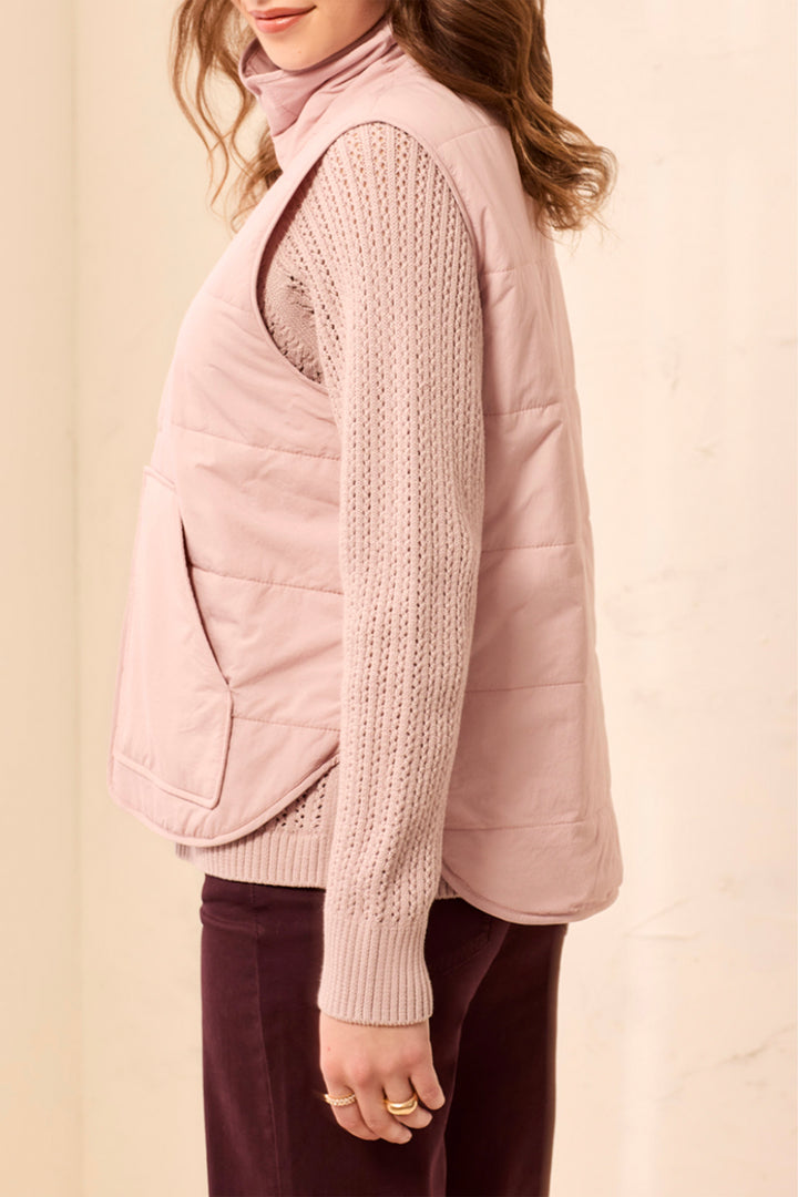 A woman shown from the side modelling a blush pink puffer vest and sweater standing in front of a light beige background. 