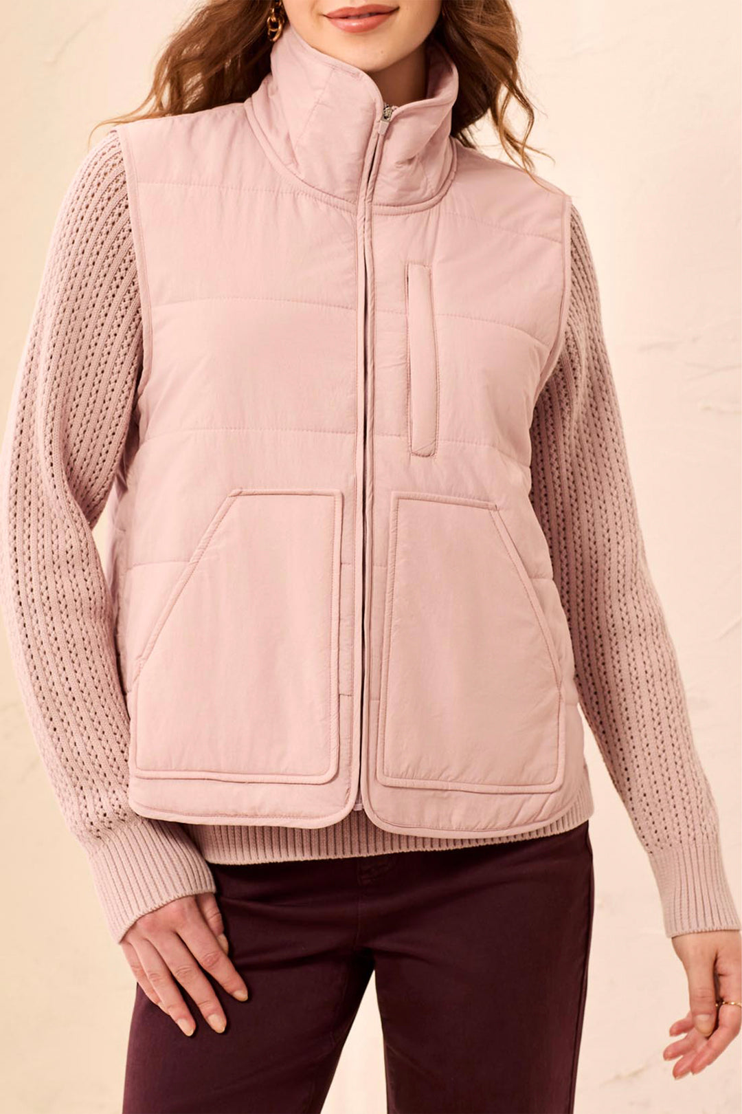 A woman modelling a blush pink puffer vest and sweater standing in front of a light beige background. 