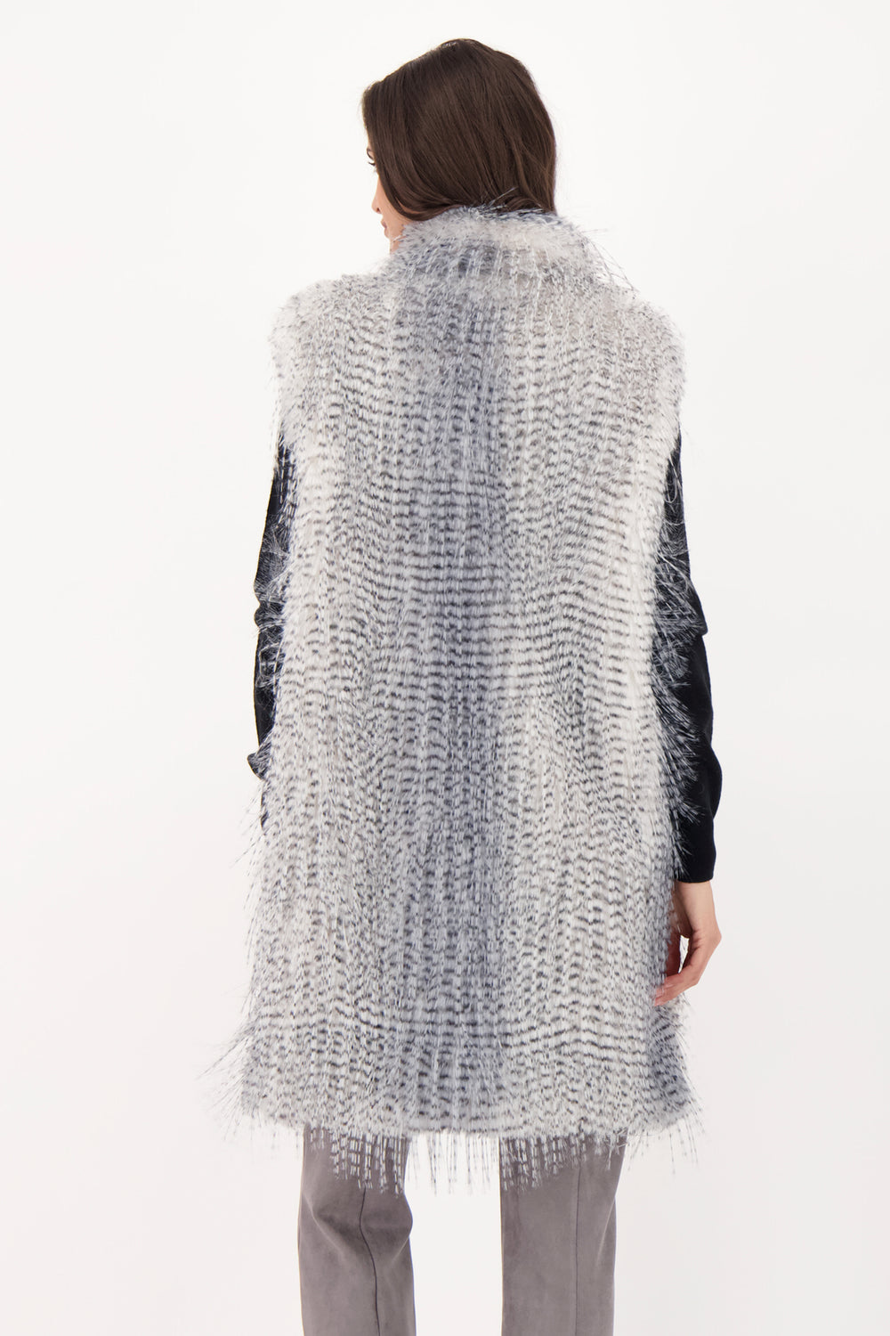 A woman shown from the back wearing a long grey faux fur vest over a black top and grey jeans. 