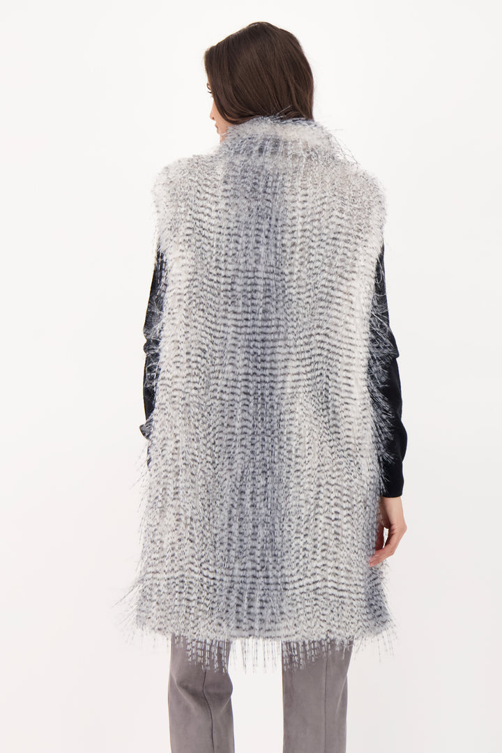 A woman shown from the back wearing a long grey faux fur vest over a black top and grey jeans. 