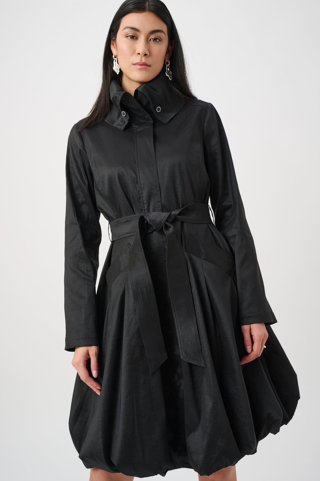A woman wearing a black dress coat with stand collar, and a balloon hem in front of a white background. 