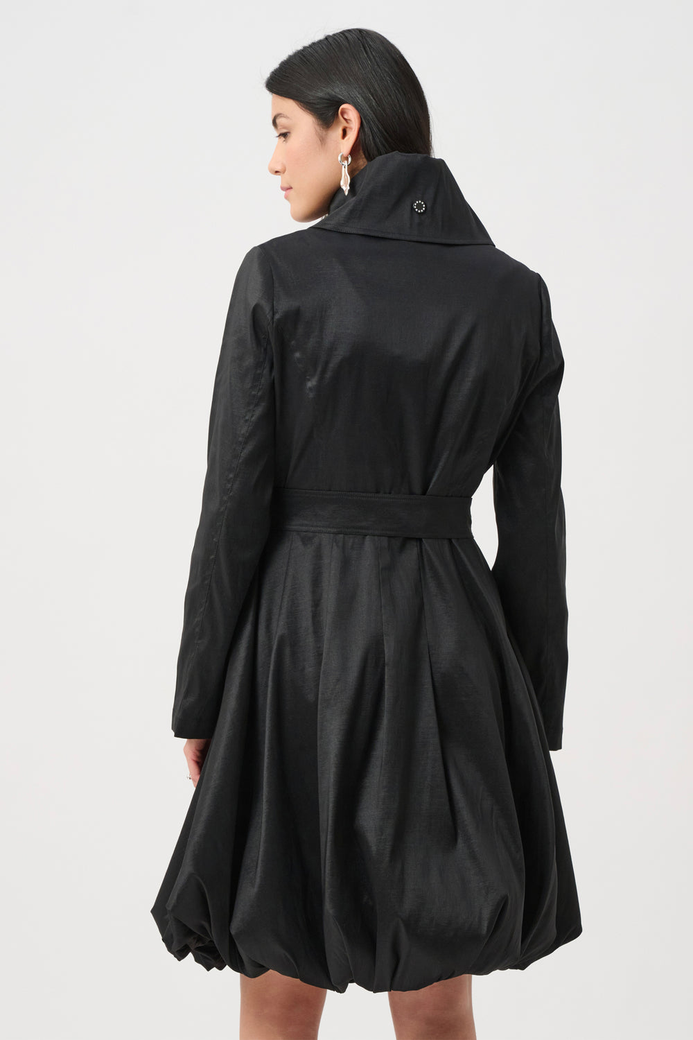 A woman shown from the back wearing a black dress coat with stand collar, and a balloon hem in front of a white background. 