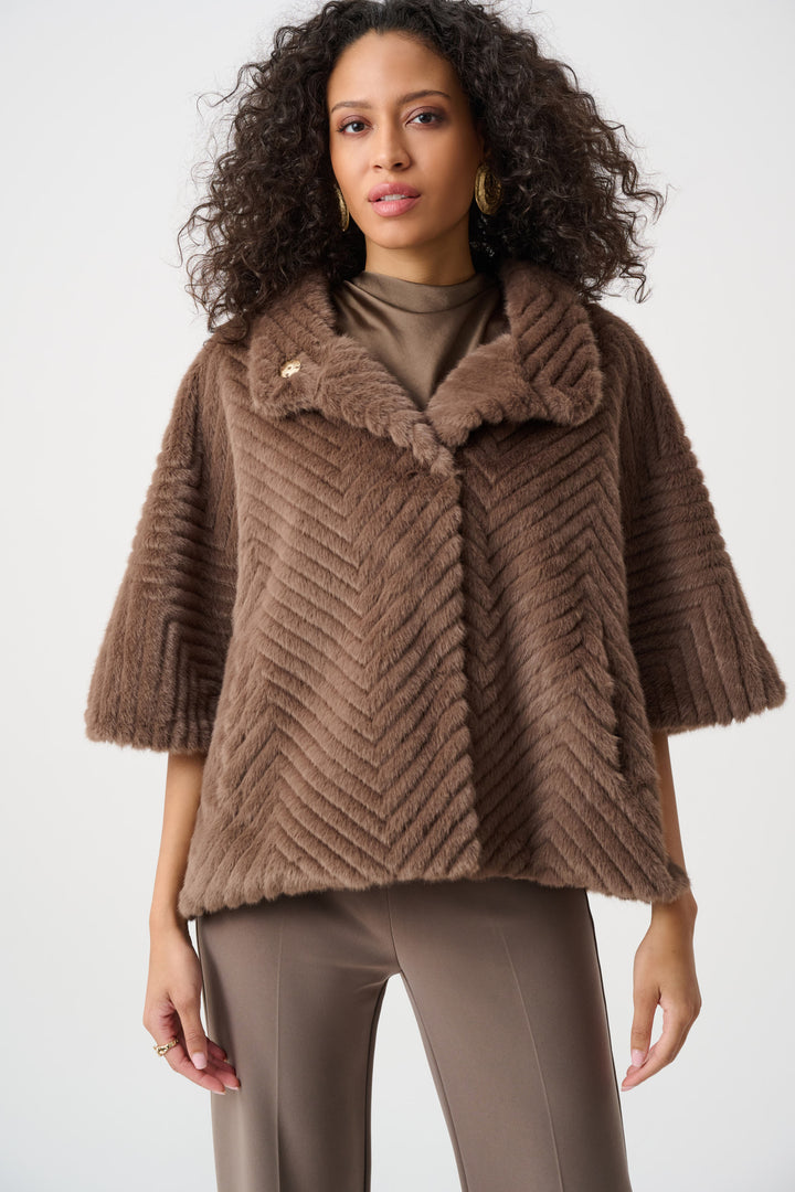 A woman wearing a brown faux fur cape jacket and brown pants standing in front of a light grey background. 
