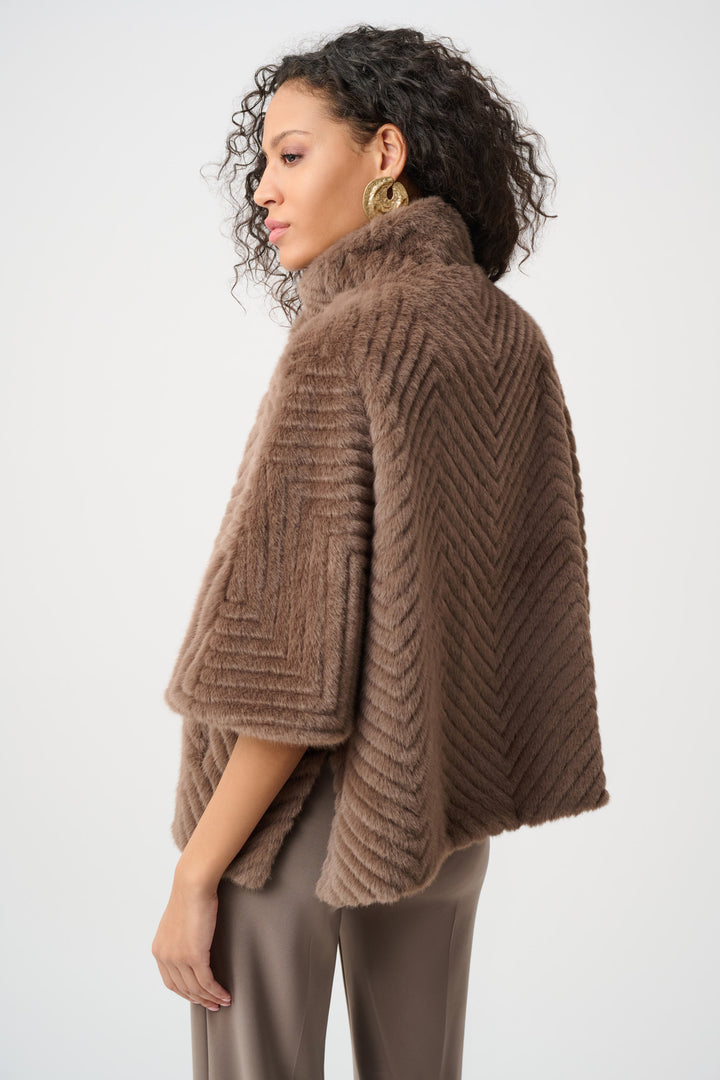 A woman shown from the back wearing a brown faux fur cape jacket and brown pants standing in front of a light grey background. 