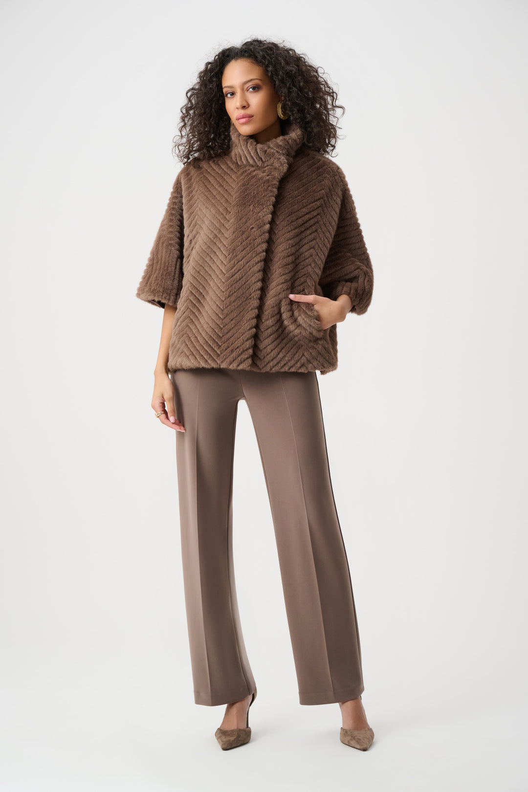A woman wearing a brown faux fur cape jacket and brown pants standing in front of a light grey background. 