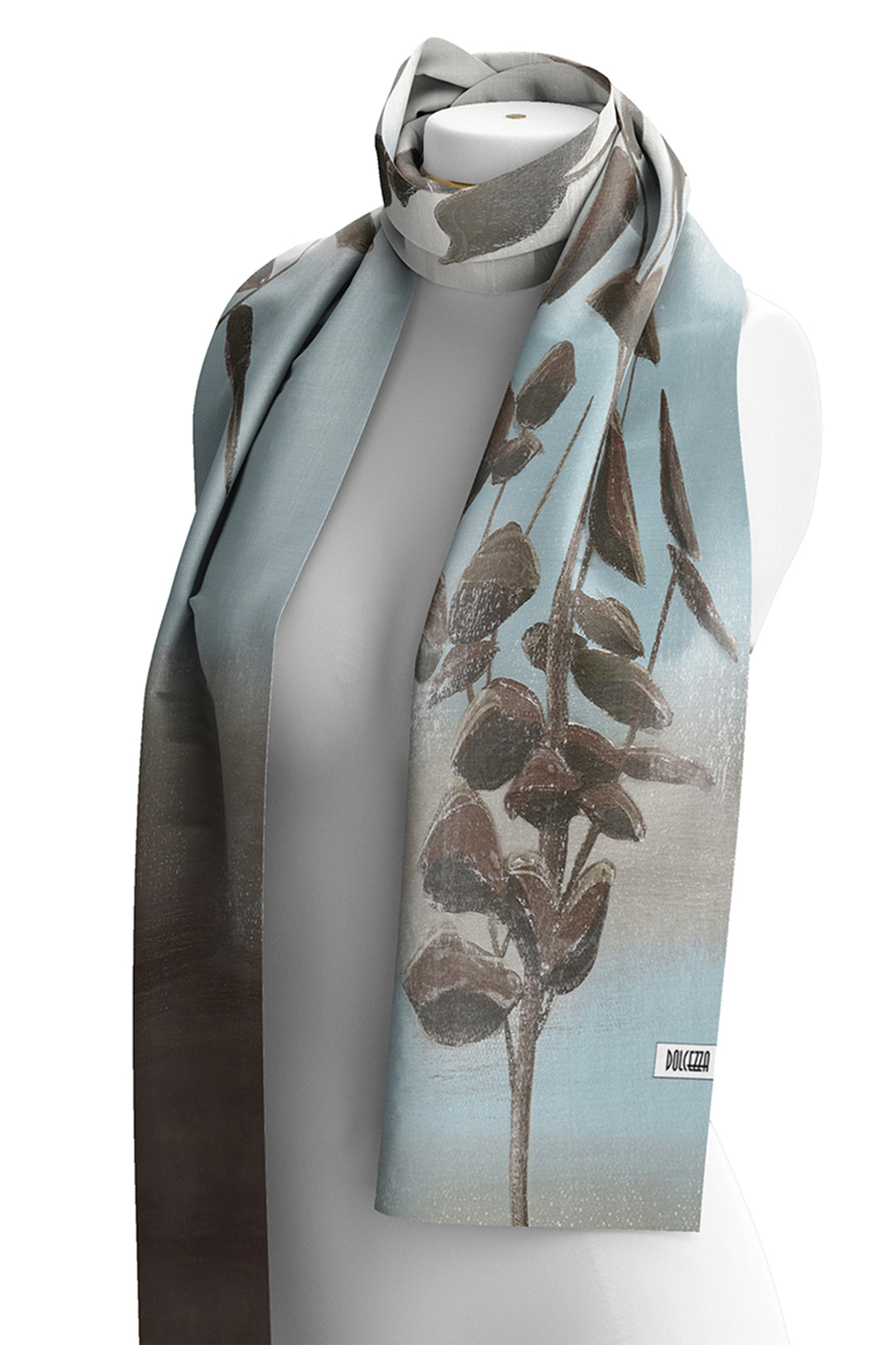 A long scarf draped on a white mannequin torso against a white background. The scarf features a botanical print of brown leaves on a light blue and gray gradient base, with a small white brand label near the lower right edge.